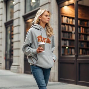 May include: A gray hoodie with the word "ACOTAR" in orange and yellow lettering. The hoodie has a drawstring hood and a front pocket. The person is wearing blue jeans and holding a coffee cup.