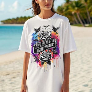 Puede incluir: Camiseta blanca con un diseño gráfico que presenta rosas negras, pájaros y el texto "YOU HAD ME AT TRIGGER WARNING". El diseño incluye un efecto de salpicadura de acuarela colorida.