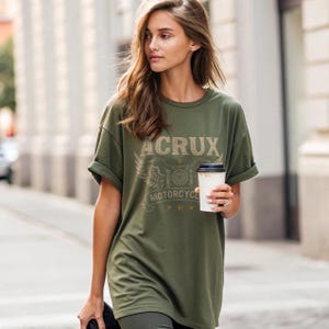 May include: An olive green short-sleeve t-shirt featuring a graphic print with the text ACRUX MOTORCYCLE, stylized wings, and three small stars. The shirt has a crew neckline and rolled sleeves, providing a relaxed fit for casual daily wear.