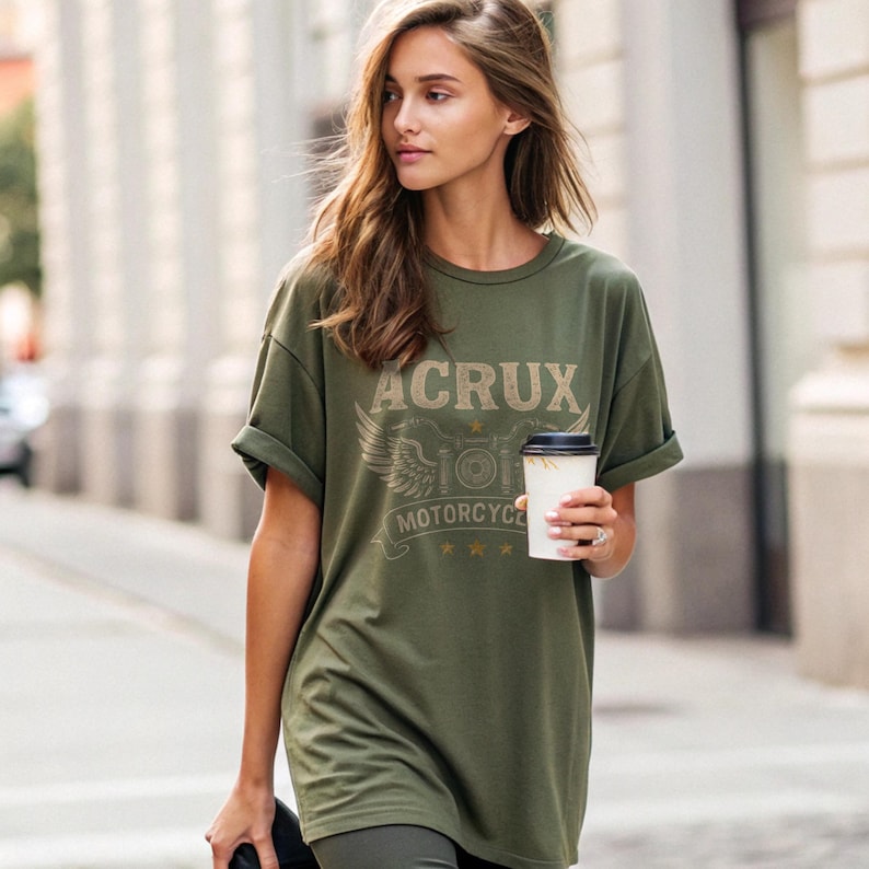 May include: An olive green short-sleeve t-shirt featuring a graphic print with the text ACRUX MOTORCYCLE, stylized wings, and three small stars. The shirt has a crew neckline and rolled sleeves, providing a relaxed fit for casual daily wear.