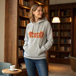 May include: A gray hooded sweatshirt with the word "ACOTAR" in a red and yellow design across the chest. The sweatshirt has a front pocket and is being worn with blue jeans. The background features a wooden bookshelf.