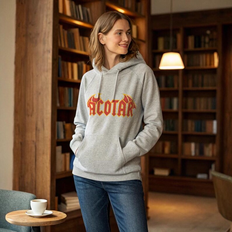 May include: A gray hooded sweatshirt with the word "ACOTAR" in a red and yellow design across the chest. The sweatshirt has a front pocket and is being worn with blue jeans. The background features a wooden bookshelf.