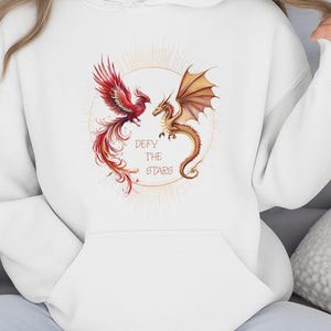 May include: White hoodie featuring a phoenix and a dragon facing each other within a golden sunburst design. The text "DEFY THE STARS" is centered below the creatures. The phoenix is red, and the dragon is gold.