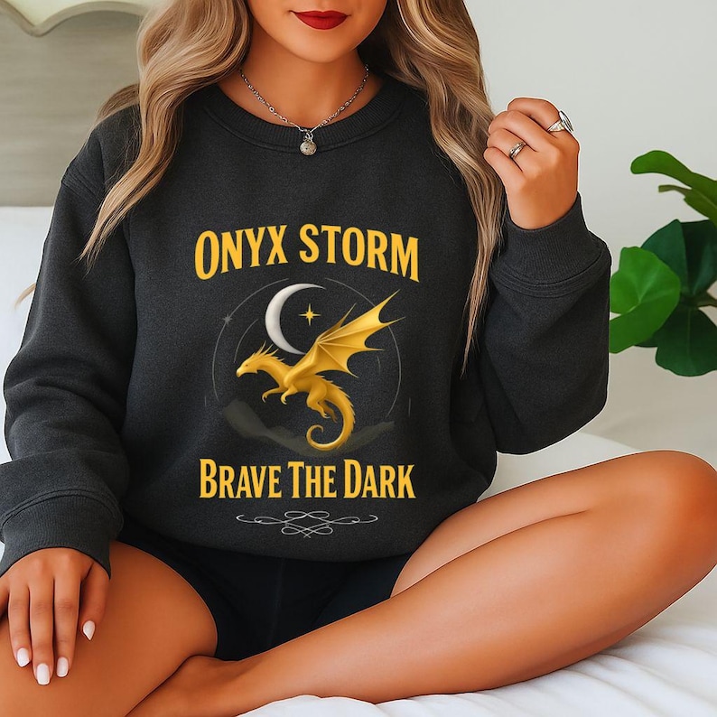 Dark charcoal gray sweatshirt featuring golden dragon graphic with crescent moon and star, and bold fantasy text reading 'ONYX STORM – BRAVE THE DARK'. Worn by model sitting cross-legged on a bed, styled with long wavy hair, red lipstick, silver pendant necklace, and gemstone ring. Cozy, mystical fashion perfect for romantasy readers, dragon lovers, and fans of bold, story-inspired apparel. Ideal gift for those who embrace magical aesthetics and dark fantasy vibes.
