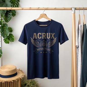 May include: A navy blue short-sleeve t-shirt featuring a graphic print on the chest. The design includes the text ACRUX in a distressed font above a stylized motorcycle emblem with wings, the word MOTORCYCLES on a banner, and three small stars.