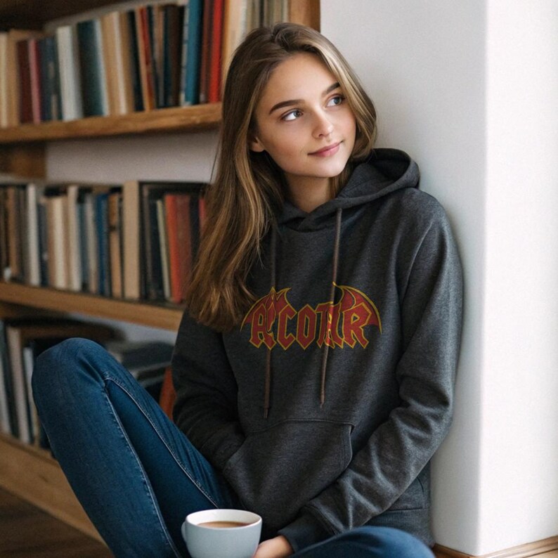 May include: Dark gray hoodie with the word "RCOTR" in a red and gold gothic font. The hoodie has a drawstring hood and a front pocket. The person is wearing blue jeans and holding a light blue mug.