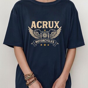 May include: A navy blue short-sleeve t-shirt featuring a graphic print in light yellow. The design includes the text ACRUX above a stylized motorcycle emblem with wings, the word MOTORCYCLES on a banner, and three small stars at the bottom.