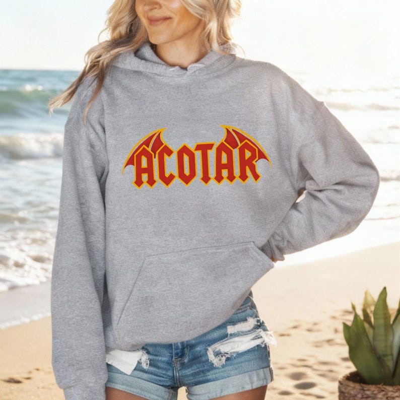 May include: A gray hoodie with the word "ACOTAR" in red, stylized text with yellow accents. The hoodie has a front pocket and a hood. The person in the image is wearing denim shorts.
