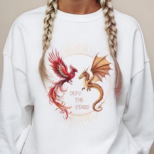May include: A white long-sleeve sweatshirt featuring a graphic print of a red phoenix and a gold dragon facing each other within a circular sunburst design. The text "DEFY THE STARS" is printed in the center between the two mythical creatures.