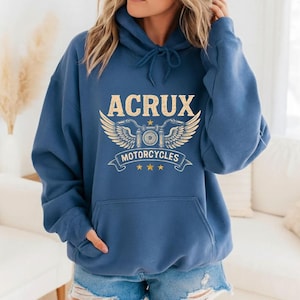 May include: A blue hooded sweatshirt featuring a graphic print in beige. The design includes the text ACRUX above an illustration of motorcycle handlebars with wings, the word MOTORCYCLES on a banner, and three small stars below.