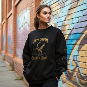 Puede incluir: Sudadera negra con el texto "ONYX STORM BRAVE THE DARK" en letras doradas. El dise&ntilde;o incluye una luna creciente, estrellas y un drag&oacute;n. La sudadera tiene cuello redondo y mangas largas.