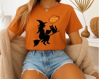 Op de afbeelding: Een oranje T-shirt met korte mouwen en een zwarte afbeelding van een heks op een bezemsteel die een boek voorleest aan een kat. Een tekstballon boven de kat bevat de tekst "IS THIS SHIT FUR REAL" in oranje letters op een witte achtergrond.