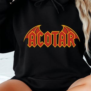 May include: Black hoodie with the word "ACOTAR" in red and yellow lettering. The design features a stylized font with wing-like accents above the text. The hoodie has a drawstring hood and long sleeves.
