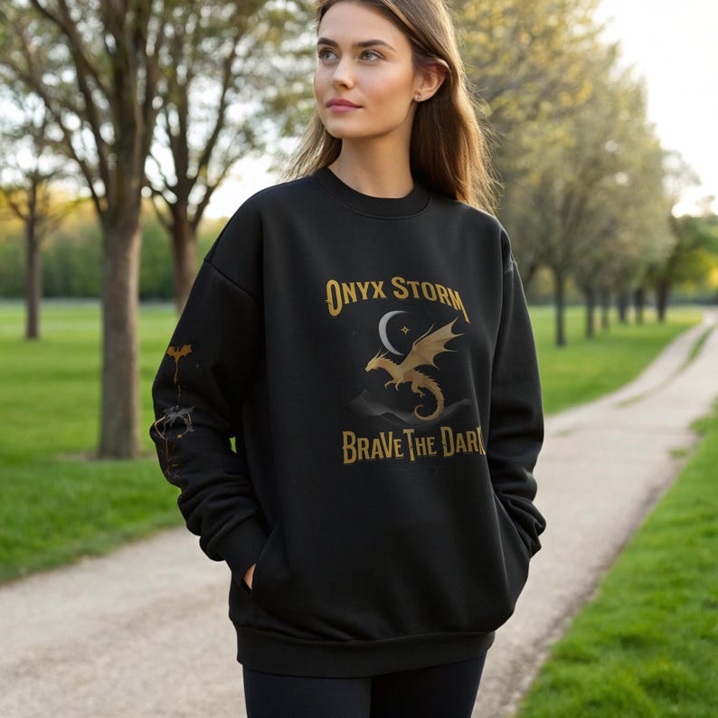 Puede incluir: Sudadera negra con el texto "ONYX STORM" sobre un gr&aacute;fico de drag&oacute;n dorado y las palabras "BRAVE THE DARK". La sudadera tiene cuello redondo y mangas largas. La persona lleva leggings negros.