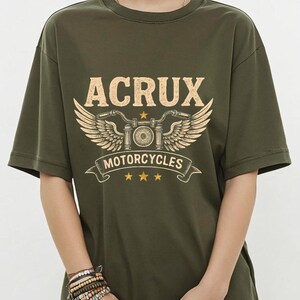 May include: An olive green short-sleeve t-shirt featuring a graphic print in light yellow. The design includes the text ACRUX, a stylized motorcycle handlebar with wings, the word MOTORCYCLES on a banner, and three small stars below the banner.