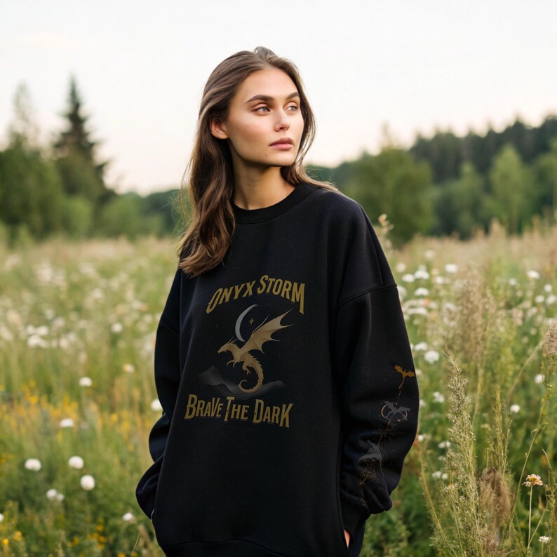 Puede incluir: Sudadera negra con el texto "ONYX STORM" sobre un gr&aacute;fico de drag&oacute;n y las palabras "BRAVE THE DARK". La sudadera tiene cuello redondo y mangas largas. La imagen est&aacute; tomada al aire libre en un campo de flores silvestres.