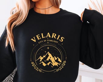 Puede incluir: Sudadera negra con texto y gráfico dorados. El texto dice "VELARIS CITY OF STARLIGHT" sobre un diseño de cordillera. El diseño está encerrado en un círculo con estrellas y hojas de laurel. La sudadera es de cuello redondo.