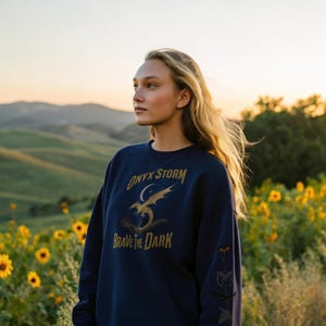 Puede incluir: Sudadera azul marino con texto dorado y un dise&ntilde;o de drag&oacute;n. El texto dice "ONYX STORM" encima de un drag&oacute;n y un dise&ntilde;o de luna creciente, con "BRAVE THE DARK" debajo. La sudadera se usa al aire libre en un campo de girasoles.