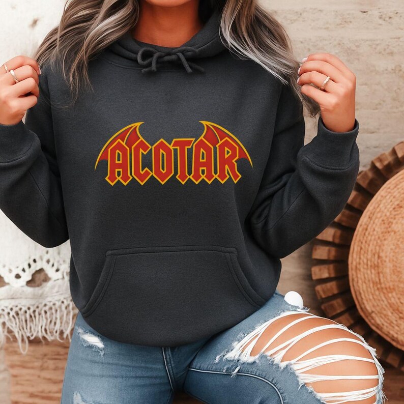 May include: Dark gray hoodie with the word "ACOTAR" in a red and yellow design. The text has a stylized, angular font with wing-like accents. The hoodie has a drawstring hood and a front pocket.