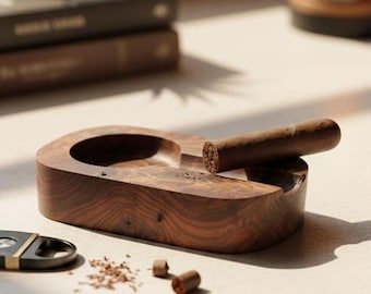 May include: A wooden ashtray with a cigar resting on it. The ashtray is oval-shaped with a deep bowl and a groove for holding the cigar. A cigar cutter and loose tobacco are also present.