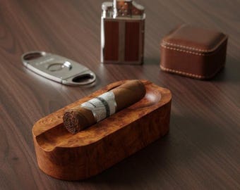 May include: A wooden cigar ashtray with a cigar, a silver cigar cutter, a brown leather lighter, and a small brown leather box. The ashtray has a natural wood grain finish.