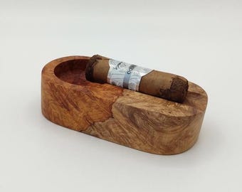 May include: A handcrafted wooden cigar ashtray with a natural wood grain finish. A brown cigar with a silver and white label rests in the ashtray. The ashtray has a rounded shape with a recessed area for the cigar.