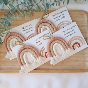 May include: Four rainbow-coloured crocheted keychains with tassels, each attached to a gold key ring. The keychains are displayed on a wooden surface with a card that reads "We are very happy because you are a part of our life."