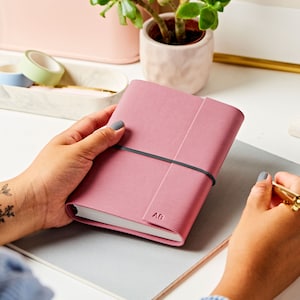 The A6 vegan leather notebook in dusky rose with a grey band around the middle. The notebook is personalised with the initials AB. The pink notebook is being held by a pair of hands at a desk.