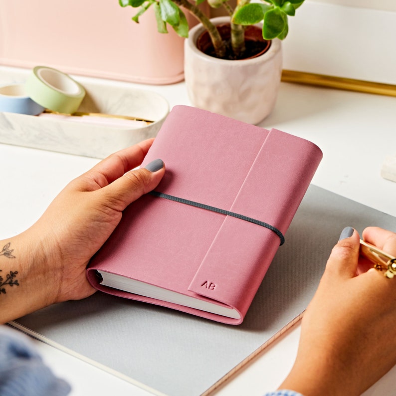 The A6 vegan leather notebook in dusky rose with a grey band around the middle. The notebook is personalised with the initials AB. The pink notebook is being held by a pair of hands at a desk.