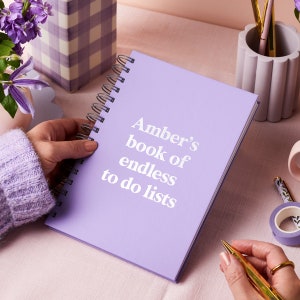 May include: A lilac spiral-bound notebook with the text "Amber's book of endless to do lists" in white. The notebook is open, and a hand is holding it. Other items on the table include a pen, washi tape, and a decorative vase with flowers.