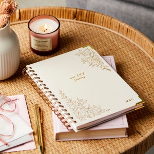 A beige IVF journal with a floral design in the top right and bottom left corners. In the centre reads the words Our IVF Journey and is personalised in the bottom right corner with the initials M&C. The IVF book is on a table.