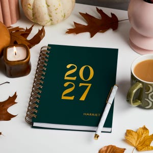 May include: A dark green spiral-bound planner with the year 2027 printed in large gold font on the front cover. The name Harriet is printed in smaller gold text below the year. A white and gray patterned pen rests diagonally across the cover.
