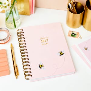 Op de afbeelding: Een lichtroze dagboek met een gouden spiraalbinding. De kaft bevat twee geïllustreerde bijen met stippellijnen als vliegroutes en de gouden tekst Claire's 2027 DIARY. Het dagboek ligt op een witte ondergrond naast een gouden pen.