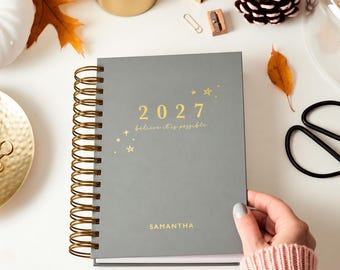 Könnte beinhalten: Ein grauer Planer mit goldener Spiralbindung. Auf dem Einband sind das Jahr 2027, der Schriftzug "believe it is possible" und der Name "SAMANTHA" in Goldfolie gedruckt, verziert mit kleinen goldenen Sternen und Funkel-Illustrationen.
