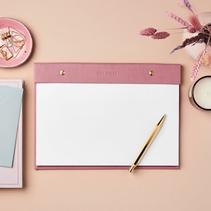 May include: A pink leather notepad with a white paper insert and a gold pen. The notepad has the text "SAM BAILEY" printed on the front.