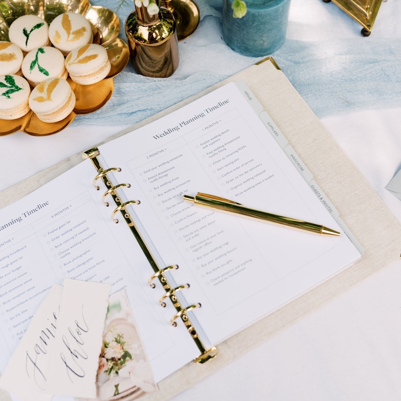 The wedding planner is laid open on a table to show a wedding planning timeline. A gold pen is laid on top of the wedding planner with two name settings and some floral macarons.