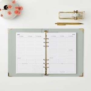 May include: An open, light green planner with gold accents, featuring a calendar for July 2025. A gold pen and tape dispenser sit nearby. A white vase with pink flowers is in the upper left corner.