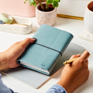 The A6 vegan leather notebook in soft teal with a grey band around the middle. The notebook is personalised with the initials CA. The teal notebook is being held by a pair of hands at a desk.