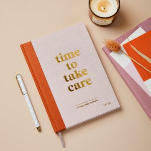 Puede incluir: Un diario de bienestar rosa claro y naranja quemado con texto dorado que dice time to take care A WELLNESS JOURNAL NINA. El diario se acompaña de un bolígrafo a cuadros blanco y azul claro, una vela encendida y elementos botánicos secos.