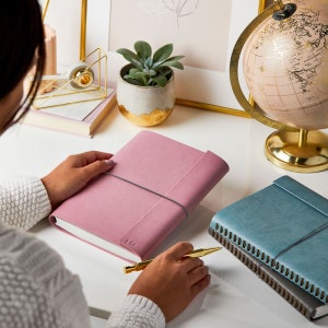 A person holding the personalised A5 vegan leather notebook in dusky pink. Next to it are two of the same notebook in soft teal and dark grey. The pink notebook is personalised in the bottom right hand corner with the initials LGF.