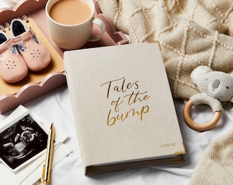 May include: A beige linen-covered baby journal with gold lettering that reads "Tales of the Bump" and the name "Jasmine". The journal is next to a pink tray with baby shoes, a mug of tea, and a baby toy.