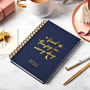 Peut inclure: Un agenda bleu foncé à reliure spirale avec une inscription en lettres dorées "find the joy in every day" et "NATALIE'S 2027 DIARY". La couverture présente de petites étoiles et un croissant de lune dorés, le tout relié par une spirale métallique.