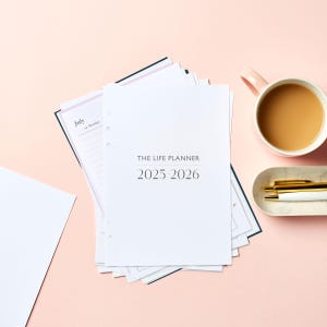 May include: A stack of white planner pages with the text "THE LIFE PLANNER 2025-2026" sits on a pink surface. A pink mug filled with a brown liquid and a small tray with two pens are also visible.