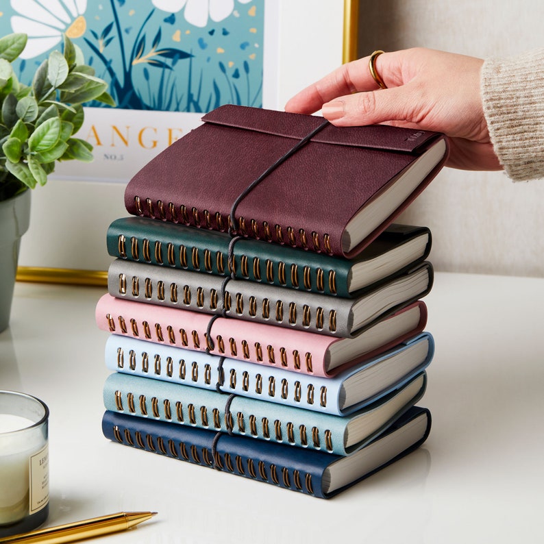 A stack showing the different colours of the A6 vegan leather notebook, with a person reaching to take the top notebook. Top to bottom, burgundy notebook, green notebook, grey notebook, pink notebook, blue notebook, teal notebook and navy notebook