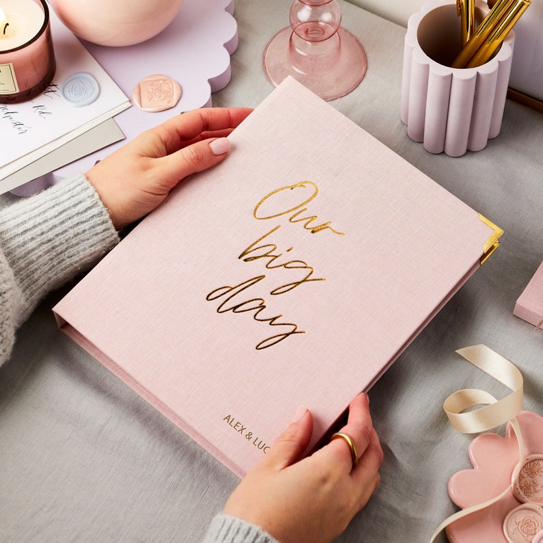 The pink wedding planner features the phrase 'our big day' in a cursive font. The names ALEX & LUCY have been embossed in gold foil on the bottom right hand side of the planner. The pink wedding planner is on a desk with wax seals.