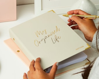 May include: A cream-colored binder with gold lettering that reads "My Organised Life" and the name "ANTONIA" on the bottom right corner. The binder is open and has white pages inside.