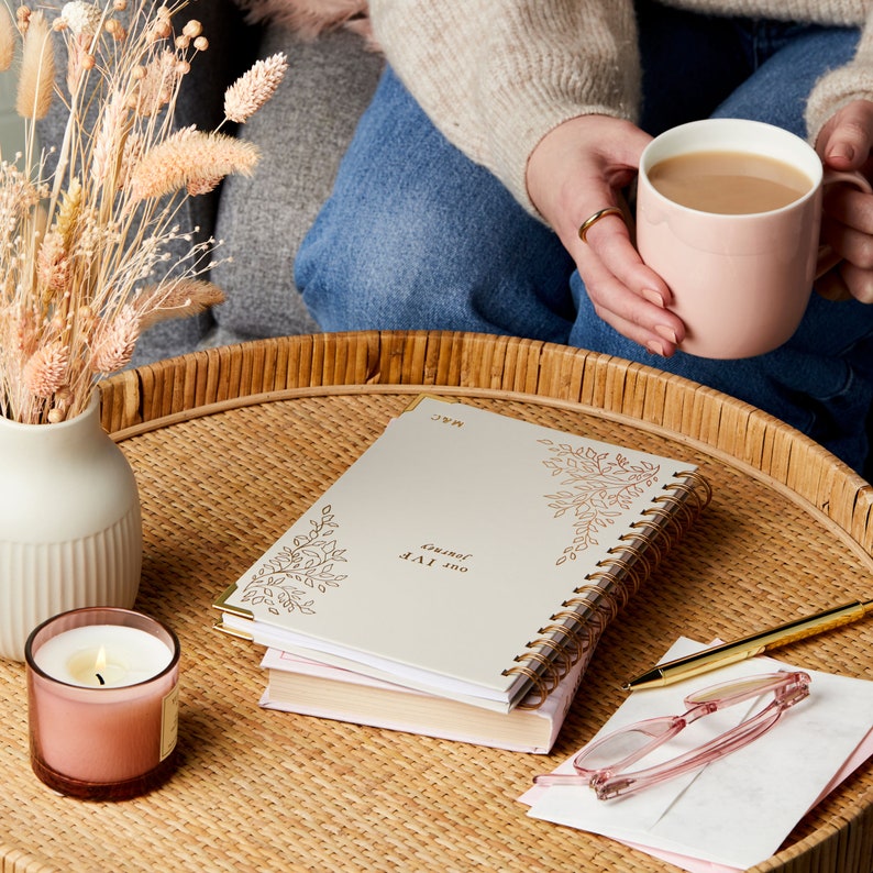 A beige IVF journal with a floral design in the top right and bottom left corners. In the centre reads the words Our IVF Journey and is personalised in the bottom right corner with the initials M&C. The IVF book is on a table.