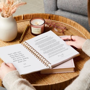 The IVF journal is being held open by a person who is looking at the first page. The first page is a quote and a note from Martha. The IVF planner is ring bound and is laid flat open on a coffee table with a candle and a pen.