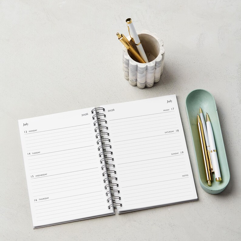 May include: An open spiral-bound planner with lined pages, dated July 13-19, 2026. A marble-look pen holder contains a gold and a white pen. A light green tray holds a gold, white, and silver pen. The items are on a light gray surface.