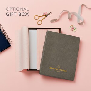 May include: A gray gift box with the text "martha brook LONDON" in gold lettering, partially open to reveal pink tissue paper and a white card. Gold scissors, a gray ribbon, and a small notebook are also visible on a pink background.